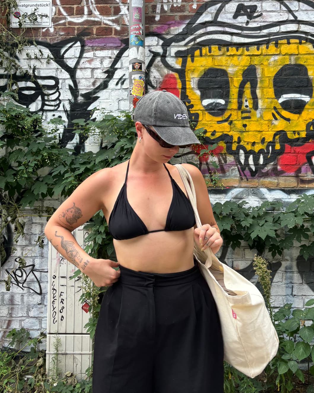 person standing with a bikini top and pants infront of a wall with graffiti, wearing the v1bes cap and sunglasses and a big tote bag