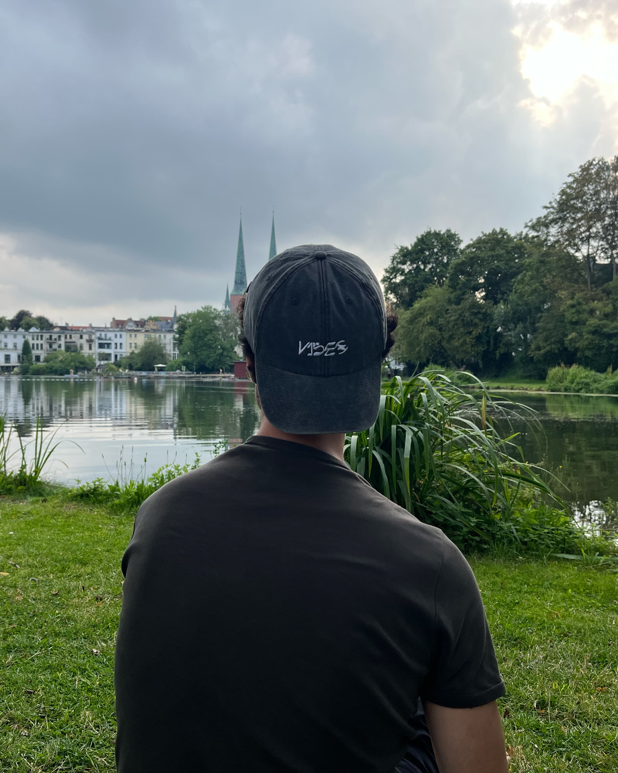 person sits with his back at a river to the camera, has the v1bes cap on